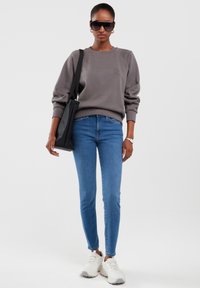 Naine kannab halli pusa, siniseid kitsaid teksapükse, valgeid tenniseid, musta käekotti, tumedaid päikeseprille ja randmekella, seistes valge tausta ees.