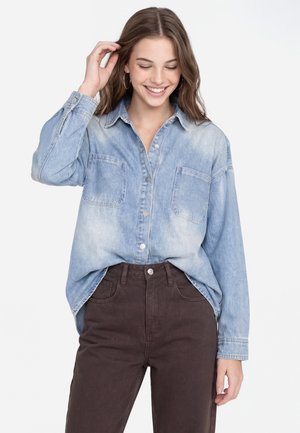 Jonge vrouw die lacht, gekleed in een lichtblauw denimoverhemd met voorzakken en donkerbruine broek, haar met één hand aanrakend.