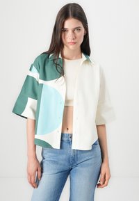 Chemise à manches courtes avec un design à demi-boutons, présentant des motifs géométriques verts et bleu clair sur un fond crème. Matériau en coton.