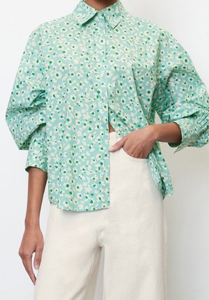 Kvinde iført en løs, mintgrøn blomsterprintet skjorte med knapper og puffede ærmer samt hvide højttaljede bukser, hånd i lommen.