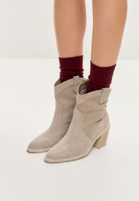 Botines de ante beige con puntera afilada y tacón ancho, que presentan detalles de corte lateral y se llevan con calcetines de canalé burdeos.