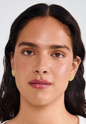 Žena s tamnom valovitom kosom nosi teksturirane zlatne spiralne naušnice i prirodni make-up s sjajnim ružičastim usnama na jednostavnoj svijetloj pozadini.