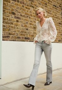 Blusa bianca in pizzo con maniche a palloncino e scollo a V, abbinata a jeans svasati grigio chiaro e stivali con tacco marrone scuro su un marciapiede testurizzato.