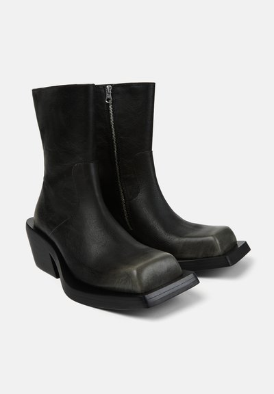 Schwarze Lederstiefel bis zur Mitte der Wade mit quadratischen Zehen, seitlichen Reißverschlüssen und einzigartig verlängerten flachen Vordersohlen auf weißem Hintergrund.