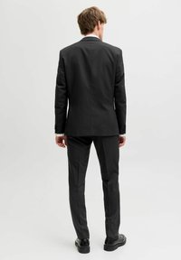 Mu�škarac stoji okrenut leđima, nosi crno odijelo s odgovarajućim hlačama i crnim cipelama na jednostavnoj svijetloj pozadini.