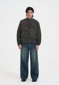 Mørkegrøn bomberjakke med broderet design, lynlås fortil og høj krave, parret med brede blå jeans og kraftige sorte sko.