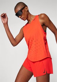 Rotes sportliches Tanktop mit perforiertem Design und passender Shorts mit gefältelten Seiten. Schwarzes Nike-Logo auf den Shorts. Elegante schwarze Sonnenbrille.