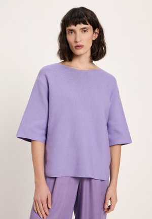 Frau mit schulterlangem dunklem Haar, die ein locker sitzendes, lavendelfarbenes Kurzarmshirt und passende Hosen trägt, vor einem schlichten Hintergrund stehend.