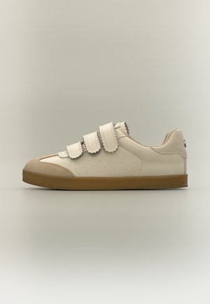 Zapatilla color crema con tres tiras de velcro con borde ondulado, suela marrón y detalles en ante en la puntera y el talón sobre fondo neutro.