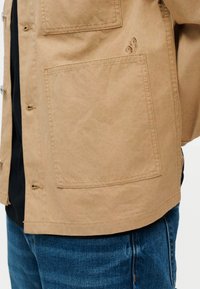 Beež jakk suure ruudukujulise eesmise taskuga, tikandid tasku lähedal, kantud musta särk ja siniste teksade peal.