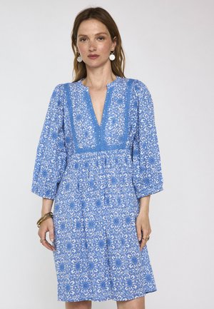 Donna che indossa un vestito con fantasia floreale blu e bianco, maniche a tre quarti, scollo a V, orecchini e braccialetti dorati.