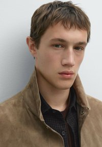 Brown suede jacket with a high collar, layered over a dark checkered shirt. Model has short hair, neutral background. Soft texture visible.