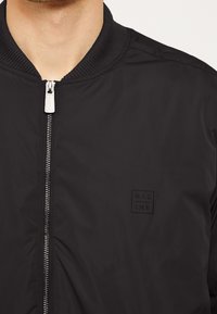 Černá bomber bunda vyrobená z hladkého materiálu. Obsahuje přední zip, žebrovaný límec a nenápadnou černou nášivku na levé straně.