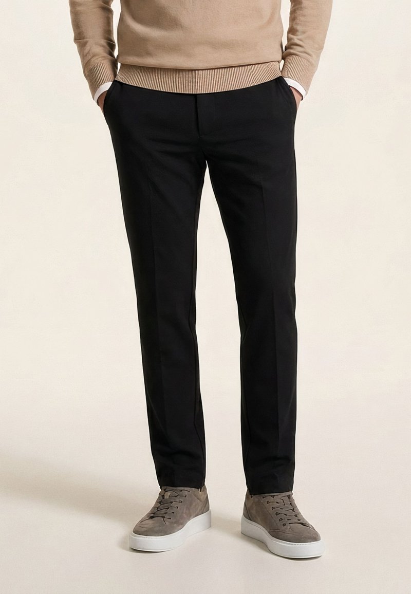 Homme portant un pantalon noir slim, un pull beige et des baskets en daim gris à semelles blanches, debout les mains dans les poches.