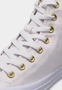Άσπρο καμβά γυναικείου ψηλού παπουτσιού με άσπρα κορδόνια περασμένα από χρυσά μεταλλικά οπές και λευκή λαστιχένια σόλα ορατή στο κάτω μέρος.