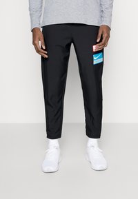 Man in zwarte taps toelopende broek met rode en blauwe patches op de dij, witte sneakers, witte sokken en een grijze lange mouwtop.