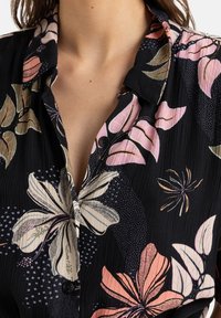 Blouse noire à motifs floraux présentant de grandes fleurs crème et rose, une texture froissée et des boutons noirs sur le devant. Col légèrement relevé.