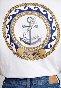 Hvid t-shirt med et blåt og gyldent maritimt design med et anker og reb, indskrevet med "MOIN, MOIN" i fed gult tekst.