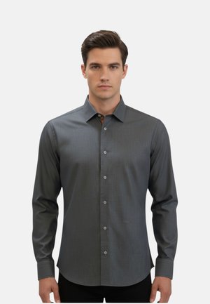 Uomo con capelli scuri che indossa una camicia grigia aderente a maniche lunghe con bottoni, in piedi davanti a uno sfondo chiaro e semplice.