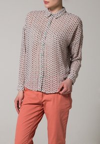 Camicia button-up con uno sfondo chiaro e un piccolo motivo di cuori colorati; maniche lunghe; colletto classico; abbinata a pantaloni arancioni.