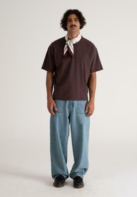 Mand med krøllet hår og overskæg, iført en mørkebrun oversized t-shirt, lyseblå bukser med vidde, sorte sko og et hvidt mønstret halstørklæde.