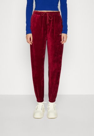 Personne portant un pantalon de jogging en velours rouge avec des chevilles élastiques, un haut bleu à manches longues et des baskets blanches à lacets, debout devant un fond blanc.