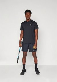 Čierne polo tričko s krátkym rukávom a detailom loga, doplnené tmavými šortkami. Model drží tenisovú raketu a loptu, na nohách má čierne tenisky.