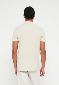 Homme portant un polo beige à manches courtes et un pantalon clair, debout de dos sur un fond blanc uni.