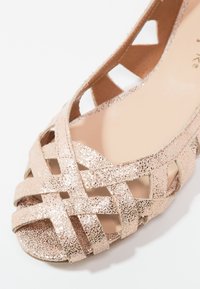 Sandale à bout ouvert avec des brides croisées scintillantes couleur or rose et une semelle intérieure beige, présentant des découpes latérales partielles pour la respirabilité.