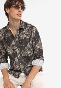 Camicia marrone a motivi con delicati disegni floreali beige chiaro. Presenta un tessuto testurizzato e polsini a quadri bianchi. Modello che indossa occhiali da sole.