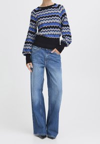 Pull-over en maille avec motif en zigzag bleu, gris et noir, associé à un jean large bleu clair et des escarpins noirs à bout pointu.