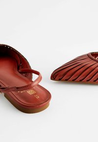 Des sandales en cuir rouge présentent un design ouvert à l'avant avec une fine lanière et des découpes diagonales. La semelle est plate et de couleur brun clair.