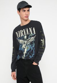 Czarna koszula z długim rękawem wykonana z bawełny, ozdobiona grafiką Nirvany z postacią z rozpostartymi skrzydłami i kolorowymi akcentami; noszona z czarnymi spodniami i czapką.