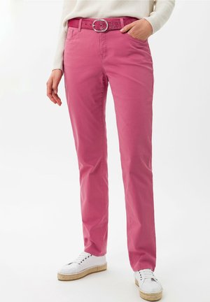 Persoon die felroze broek draagt met bijpassende glitterriem, witte top met lange mouwen en witte espadrille-sneakers, staand met hand in de zak.