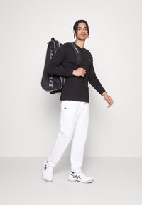 Sort langærmet skjorte, hvide sweatpants og hvide sko med sorte accenter. Modellen bærer en sort sportstaske med grå detaljer.