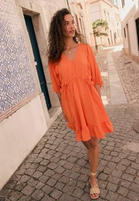Orange klänning med V-ringning, rynkad midja och volangkant. Modellen bär strappy beige sandaler mot en kullerstensgata som bakgrund.