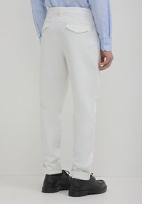 Uomo che indossa pantaloni bianchi su misura, scarpe nere stringate e una camicia a maniche lunghe di colore azzurro, in piedi contro uno sfondo bianco semplice.