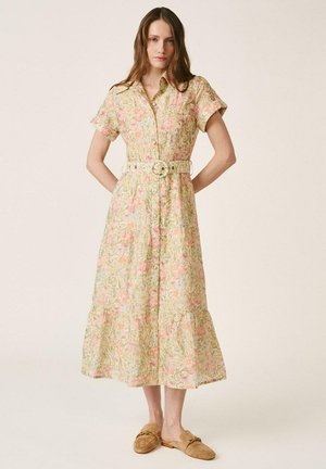 Mujer con vestido midi de flores con cinturón y mangas cortas, y mocasines marrones sin cordones, de pie con las manos detrás de la espalda.