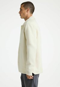 Lichtbeige, textuurjacket met een kraag, knopen aan de voorkant en zijsplitten. Heeft gestructureerde schouders en een getailleerd ontwerp.