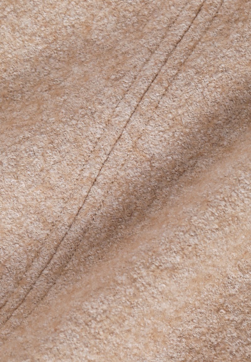 Tejido beige texturizado con una superficie suave y peluda y costuras visibles. El material parece cálido y acogedor, adecuado para prendas confortables.