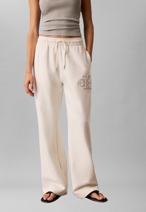 Kobieta ubrana w beżowe szerokie spodnie dresowe z logo Calvin Klein na udzie, szary krótki top i czarne klapki, stojąca na szarym tle.