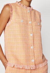 Růžová a oranžová tvídová vesta s třásněmi, má perlové knoflíky a dvě náprsní kapsy. Texturovaná tkanina s tkaným vzorem.