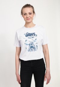 Hvid bomulds-T-shirt med et blåt billede af en haj og ordet "JAWS" i fed skrift, med sprøjtede accenter omkring designet.
