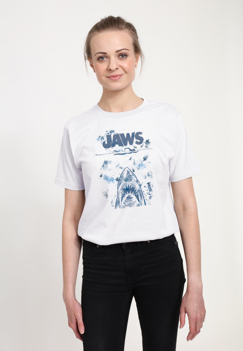 Hvid bomulds-T-shirt med et blåt billede af en haj og ordet "JAWS" i fed skrift, med sprøjtede accenter omkring designet.