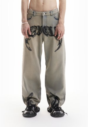 Mann trägt ausgewaschene Jeans mit weitem Bein, verziert mit schwarzer Graffiti-Schrift, und schwarze Sneaker, stehend vor weißem Hintergrund.