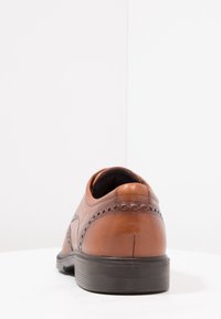 Chaussure en cuir marron avec bout arrondi, perforations décoratives et semelle en caoutchouc noire. Vue arrière montrant une finition lisse et des lignes épurées.