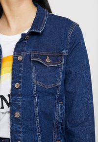 Jeansjacka i mörkblått, med klassisk krage, metallknappar, bröstficka och synlig sömnad. Materialet har en mjuk textur.