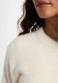 Maglione color crema, morbido al tatto, con scollatura tonda e una leggera lavorazione a coste. Il tessuto sembra soffice e caldo, adatto per il clima più fresco.