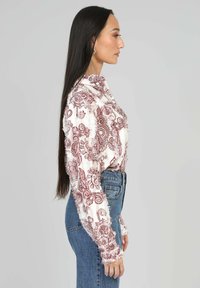 Femme aux longs cheveux raides portant un blouse à motifs floraux blancs et rouges et un jean bleu, se tenant de profil contre un fond uni.