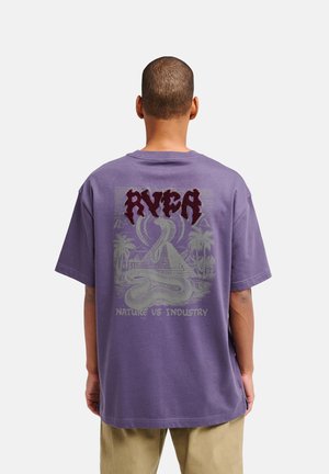Homme portant un t-shirt violet avec le texte rouge "RVCA" et un dessin de serpent sur une pyramide et des palmiers avec le texte "Nature vs Industry" au dos.